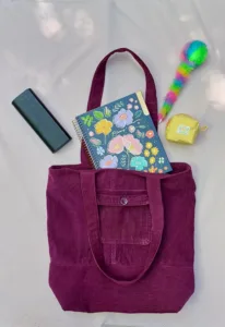 Bolso tote burdeos hecho a mano, acompañado de útiles escolares y accesorios coloridos sobre superficie clara.
